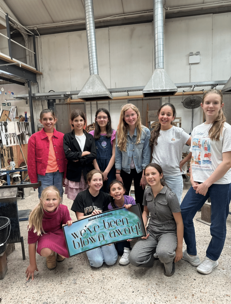 A group of kids at a glassblowing workshop holding a sign that says "We’ve been blown away!".