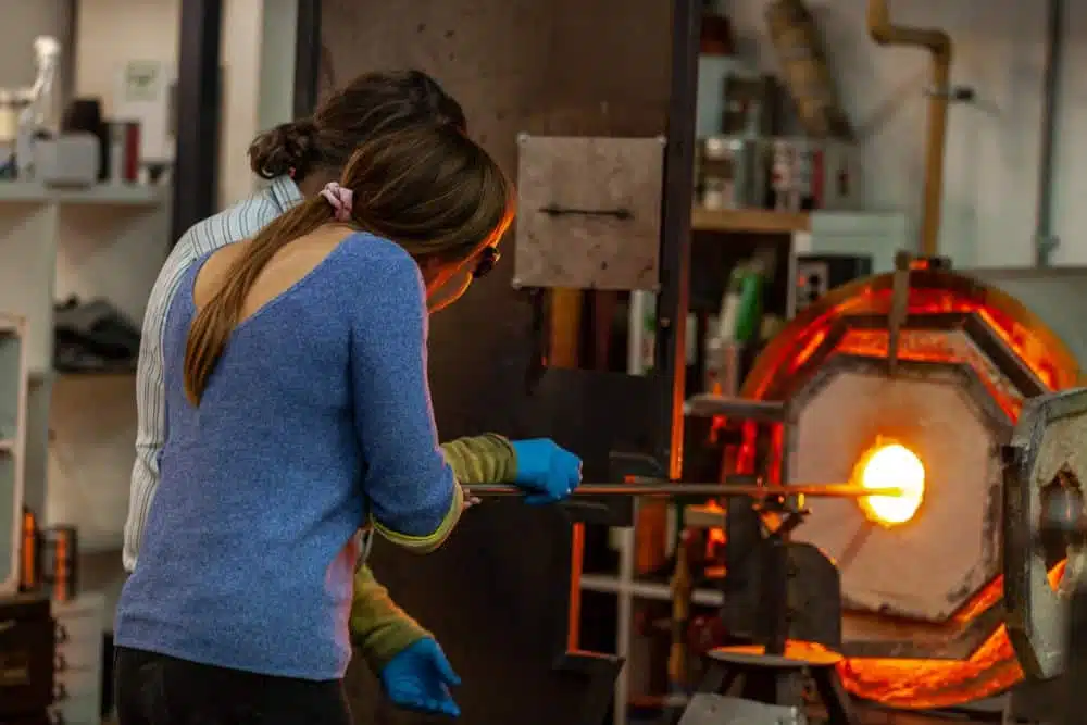 Two people are shaping glass with a rod in front of a hot furnace in a workshop.