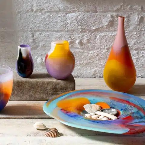 Colorful glass vases and a decorative plate with shells on a light wooden surface.