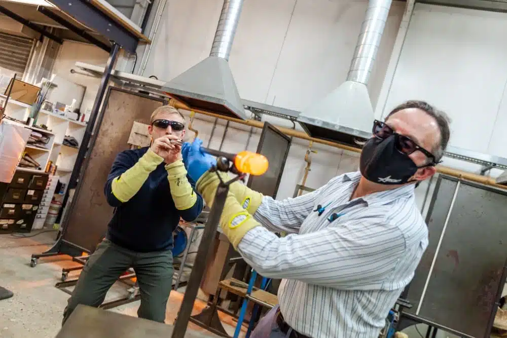 Two people wearing safety gear shape molten glass in a workshop.