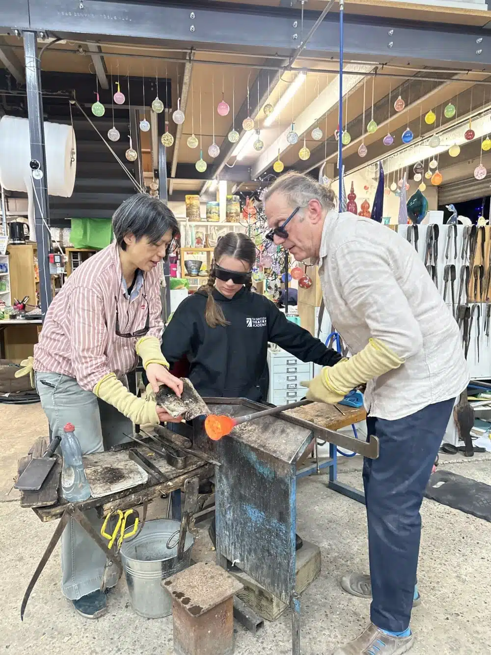 Three people working together on a glassblowing project in a colorful workshop.
