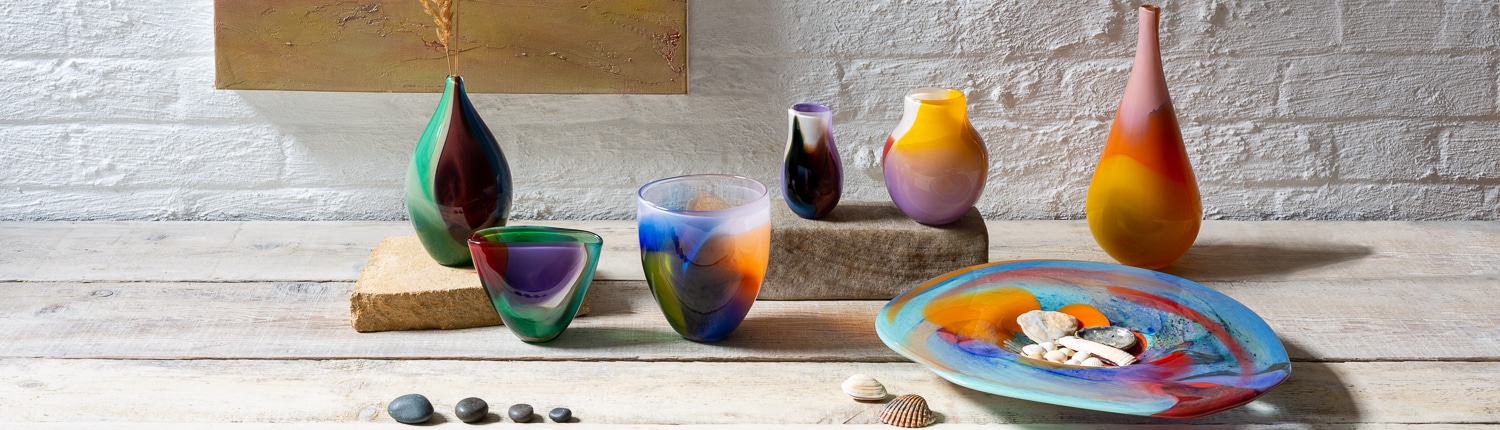 Colorful glass vases and bowls displayed on a wooden table with shells and stones.