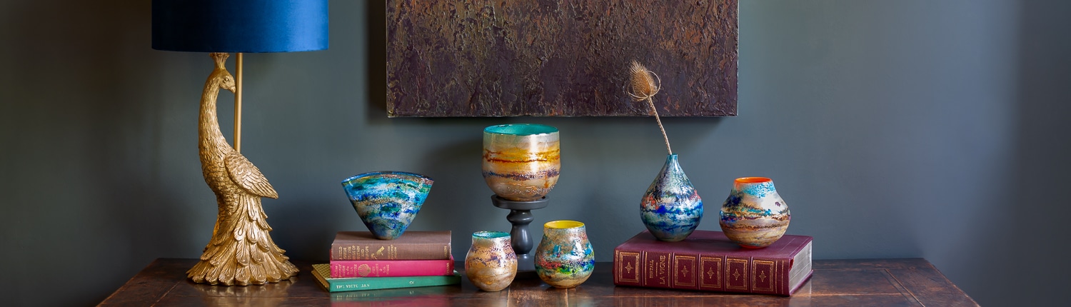 Colorful vases, books, and a gold peacock lamp are arranged on a wooden table against a gray wall.