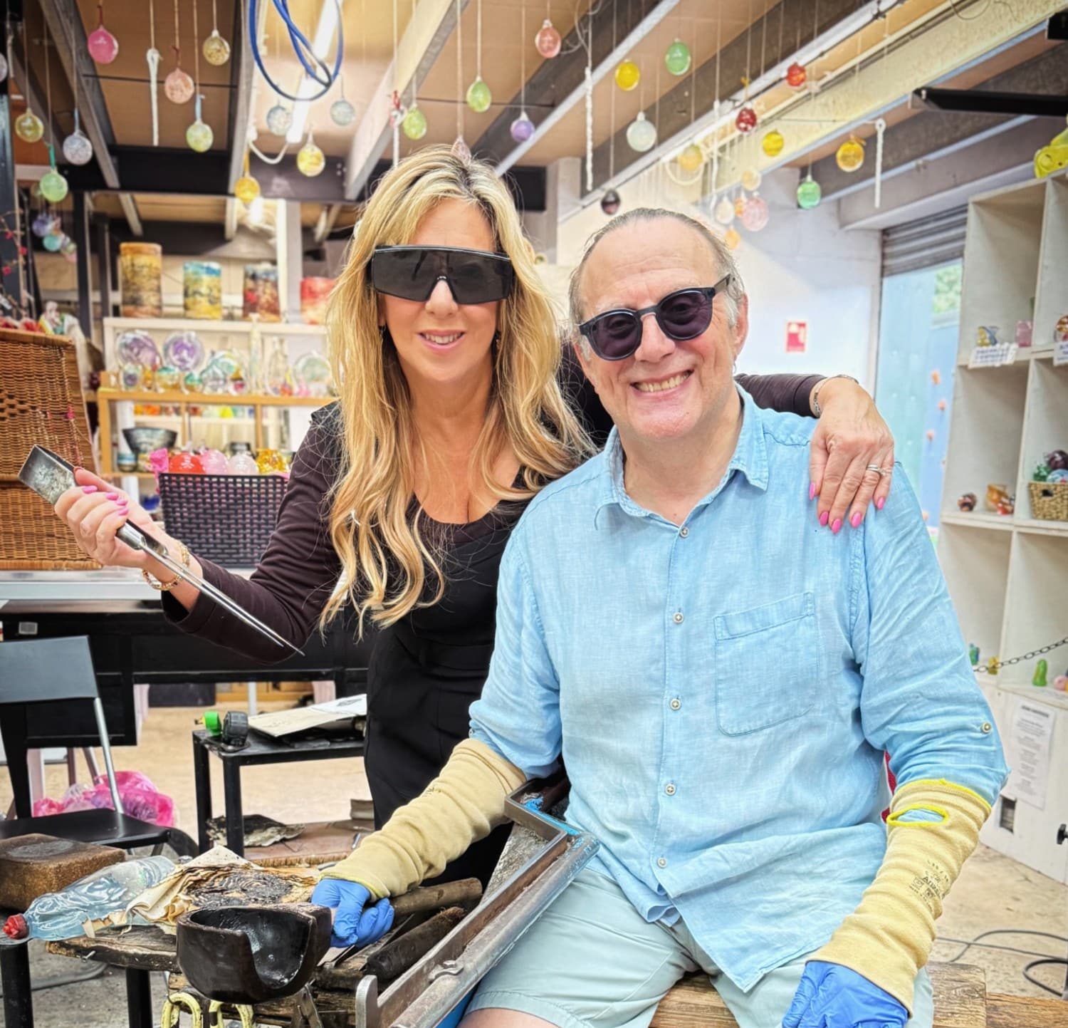 Two people smiling in a colorful glassblowing studio, wearing protective glasses and gloves.