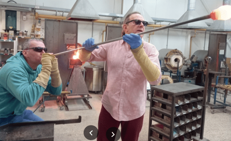 Two people blowing and shaping hot glass in a workshop, both wearing safety gloves and glasses.