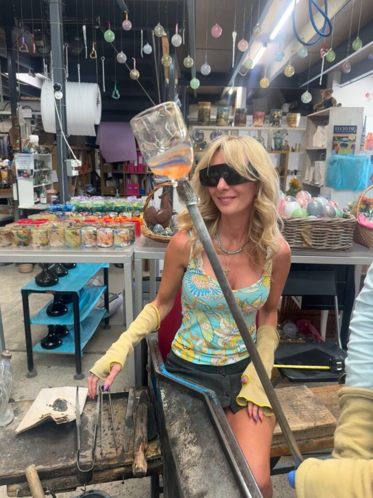 A woman with blonde hair making glass art in a workshop, wearing safety glasses and gloves.