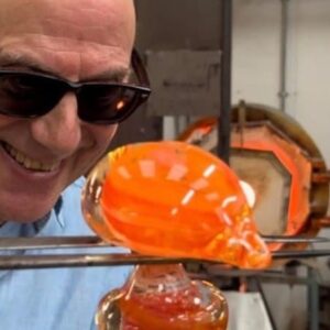 Man wearing sunglasses smiles while shaping molten glass with tools in a workshop.