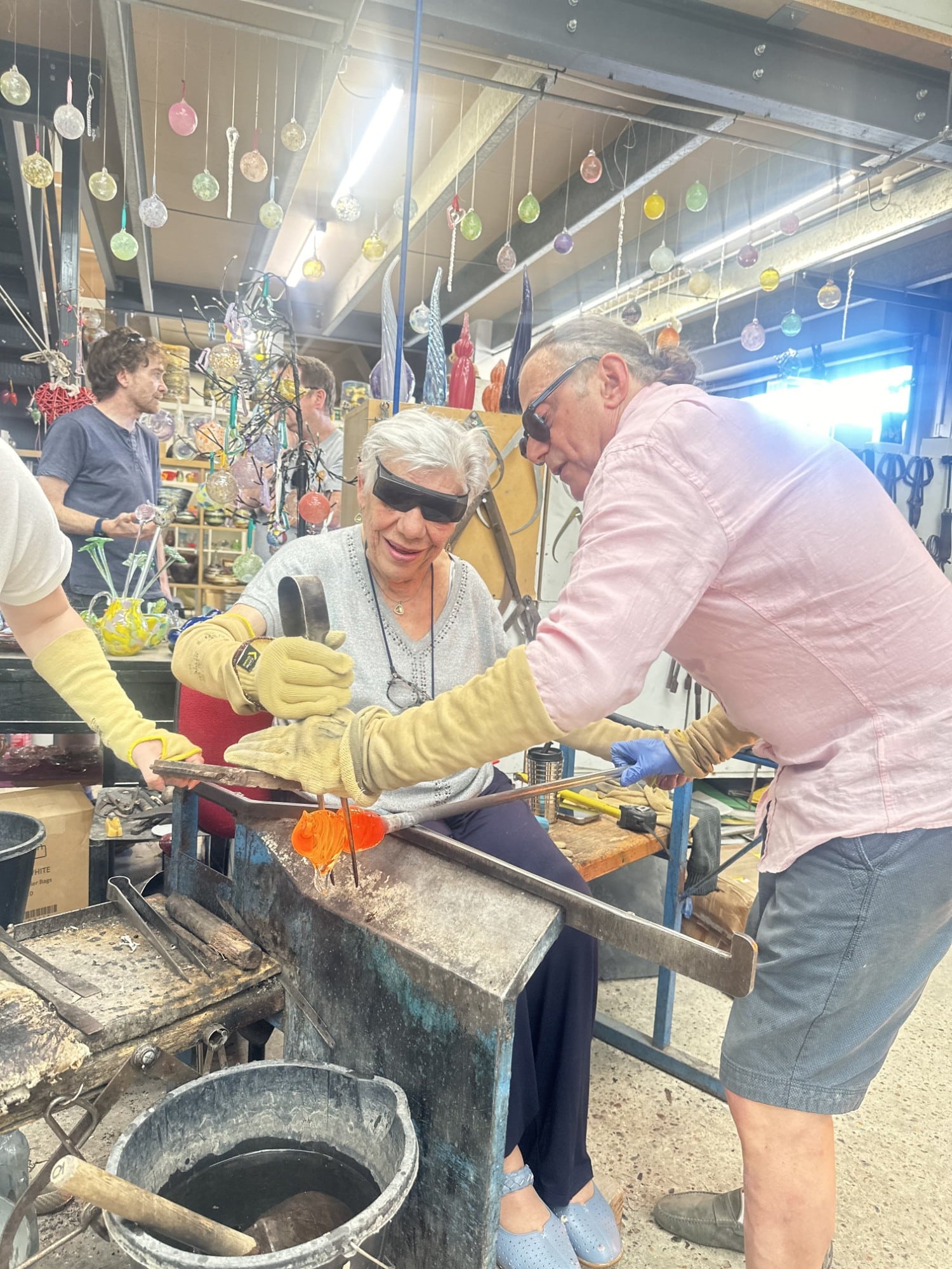 GlassblowingTogether Two people wearing safety gear work together on a glassblowing project in a colorful workshop.