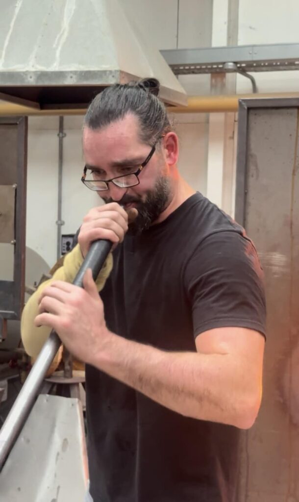 A man blowing glass in a workshop, holding a blowpipe and wearing safety glasses.
